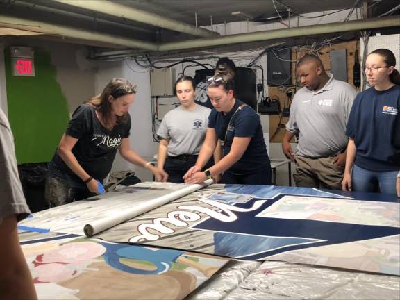 Members of the Newton First Aid Squad help unroll a section of the mural to be painted Sunday, July 30. At left is artist Caren Olmsted, who designed the mural. (Photo by Kathy Shwiff)